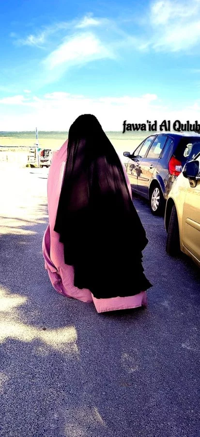 Femme de dos en jilbab rose et cape noire, debout devant l’océan sous un ciel bleu clair.