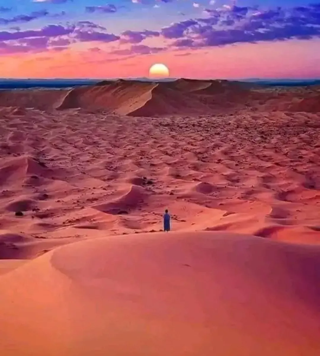 Un homme en tenue traditionnelle bleue se tient seul au sommet d’une dune, contemplant un coucher de soleil lumineux sur un paysage désertique violet-orangé, symbolisant la quête spirituelle et la guidance divine.