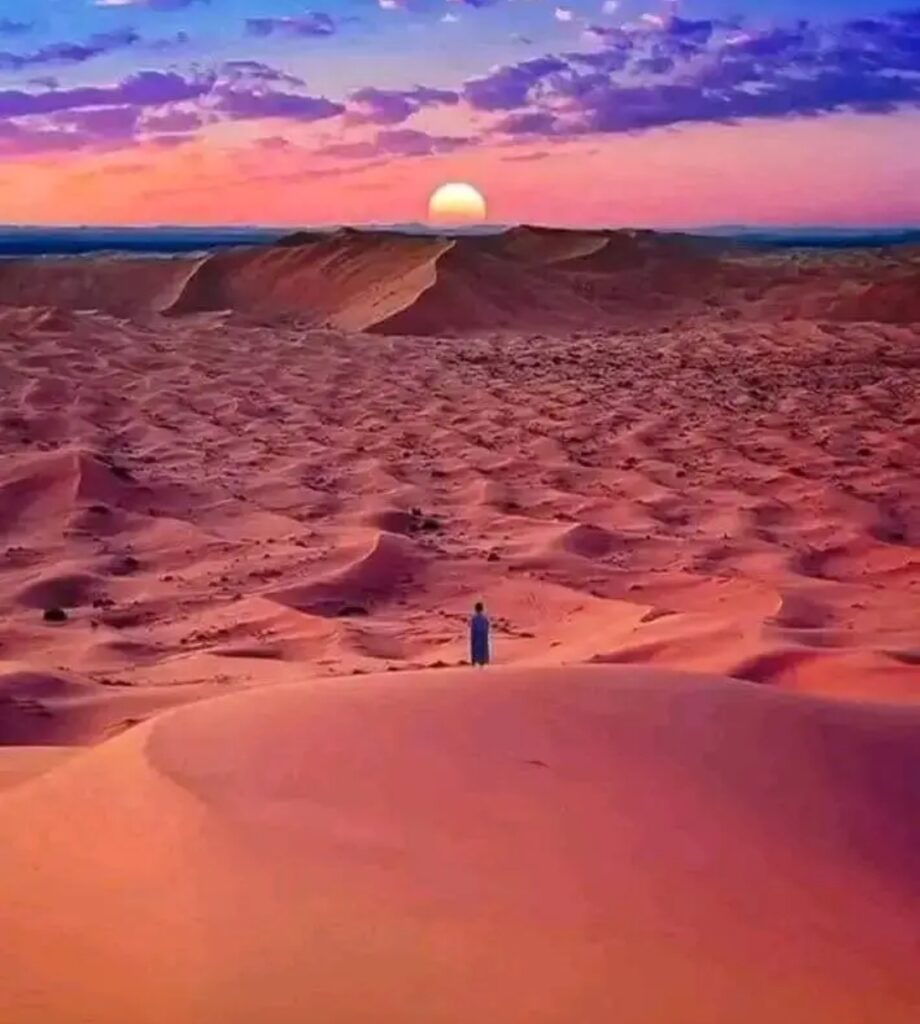 Un homme en tenue traditionnelle bleue se tient seul au sommet d’une dune, contemplant un coucher de soleil lumineux sur un paysage désertique violet-orangé, symbolisant la quête spirituelle et la guidance divine.