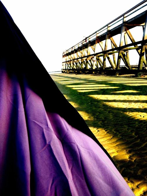 femme en jilbeb mauve et noir, debout face à la mer. siys un vieux ponton en bois, contemplant l'horizon dans un moment de receuillement spirituel. Elle exprime une profonde paix intérieur symbolisant le lacher prise et la confiance en Allah, loin du regard des gens