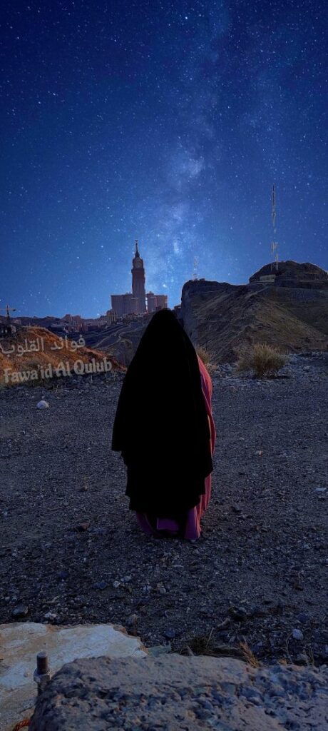 Femme seule, vêtue d’un voile noir et d’un jilbab mauve, debout au sommet d’une colline rocheuse, faisant face aux tours illuminées de La Mecque. Derrière elle, un ciel nocturne majestueux et étoilé, traversé par la Voie lactée. L’image symbolise la quête intérieure, la contemplation spirituelle et l’élévation du cœur entre le monde terrestre et le divin.